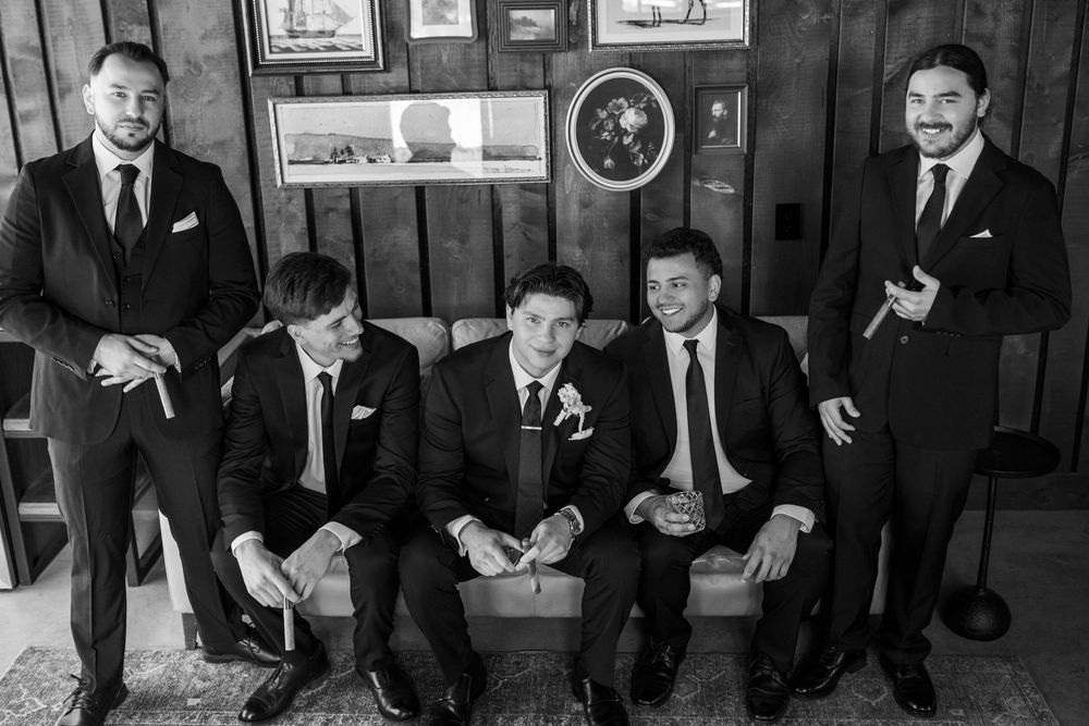 Five groomsmen in dark suits pose together in a rustic wood-paneled room, three seated on a couch and two standing, holding cigars.