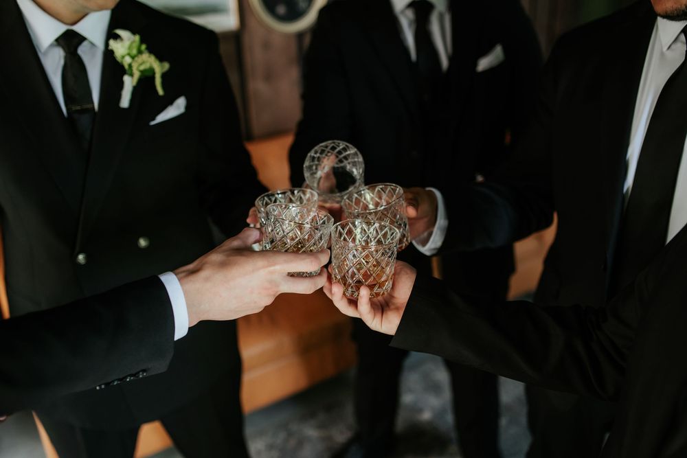 Groomsmen in dark suits clink crystal whiskey glasses together in a toast, with one wearing a white floral boutonniere.