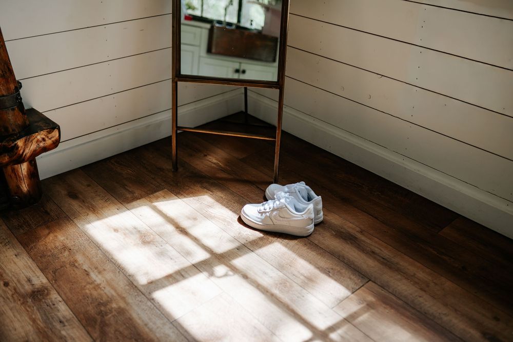 White sneakers rest on a sunlit hardwood floor beside a standing mirror in a rustic room with white wood plank walls.