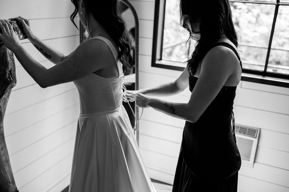 Black and white photo of a bridesmaid in a dark dress lacing the back of a bride's gown beside a bright window.