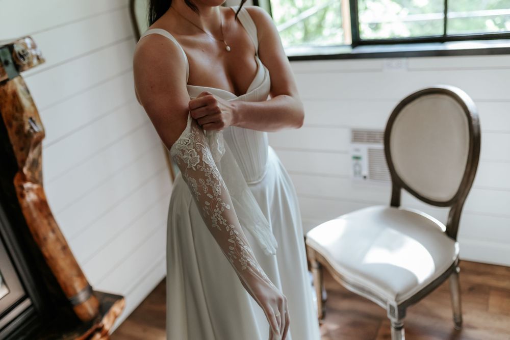 Bride in a flowing white dress pulls on a long lace glove beside an upholstered wooden chair in a bright, rustic room.