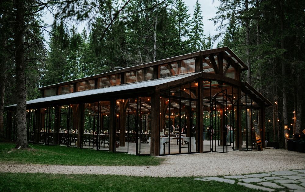 Glass-walled wedding pavilion with a timber frame and string lights, surrounded by tall evergreen trees at dusk with a gravel path in front.