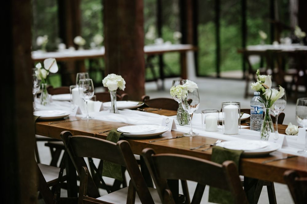 Rustic wooden banquet table with dark chairs, white floral centerpieces, place settings and glassware in a bright garden wedding venue.