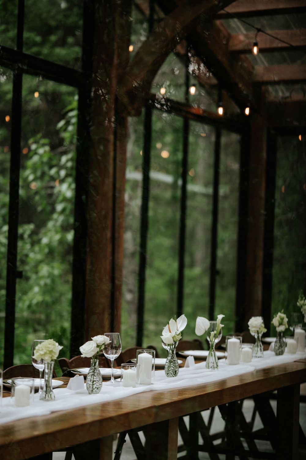 Rustic wooden dining table with white flowers in bud vases and candles inside a glass-walled forest venue with string lights.
