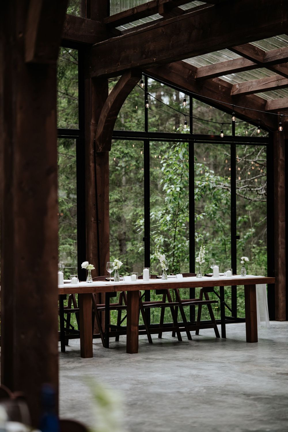Rustic wooden event pavilion with a long farm table, simple centerpieces, and floor-to-ceiling windows overlooking a lush green forest.