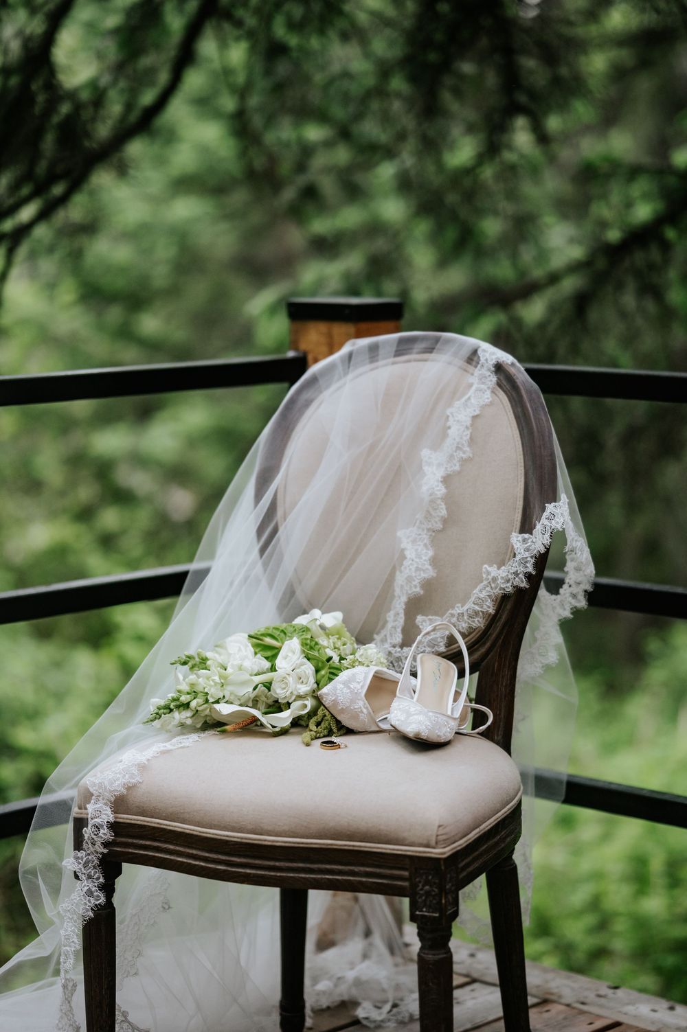 Bridal veil with lace trim draped over a vintage chair beside a bouquet and white heels on a wooden deck.