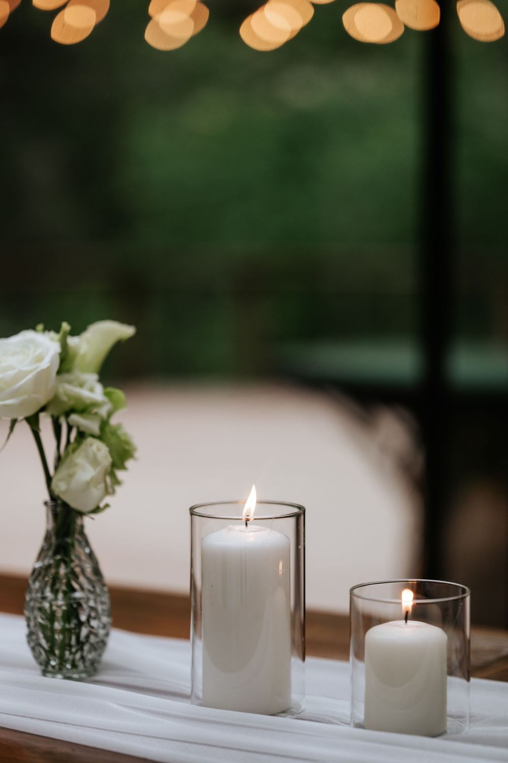 Two lit white pillar candles in glass holders beside a vase of white roses on a table with a white runner and warm string lights.