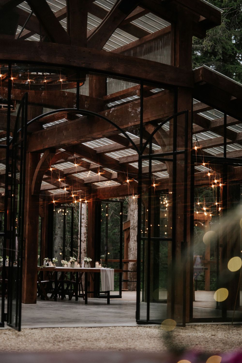 Rustic timber-frame pavilion with glass walls, string lights and a long reception table set for an elegant forest wedding dinner.