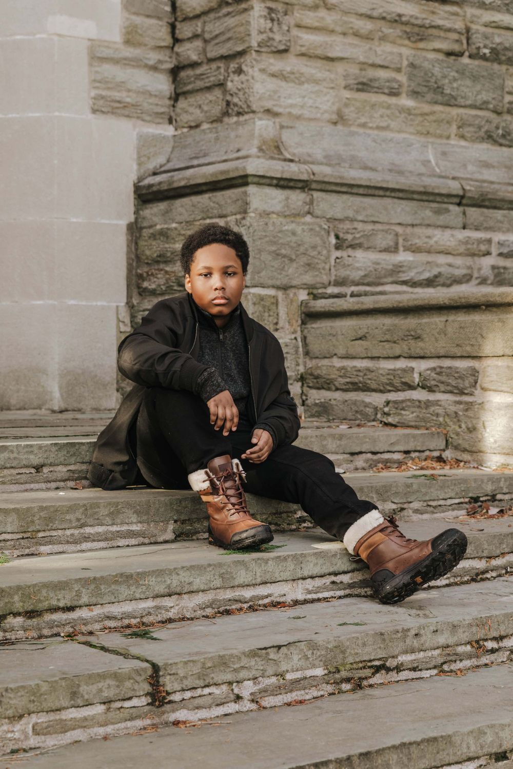 Person in black hoodie and brown boots sitting on stone steps with serious expression.