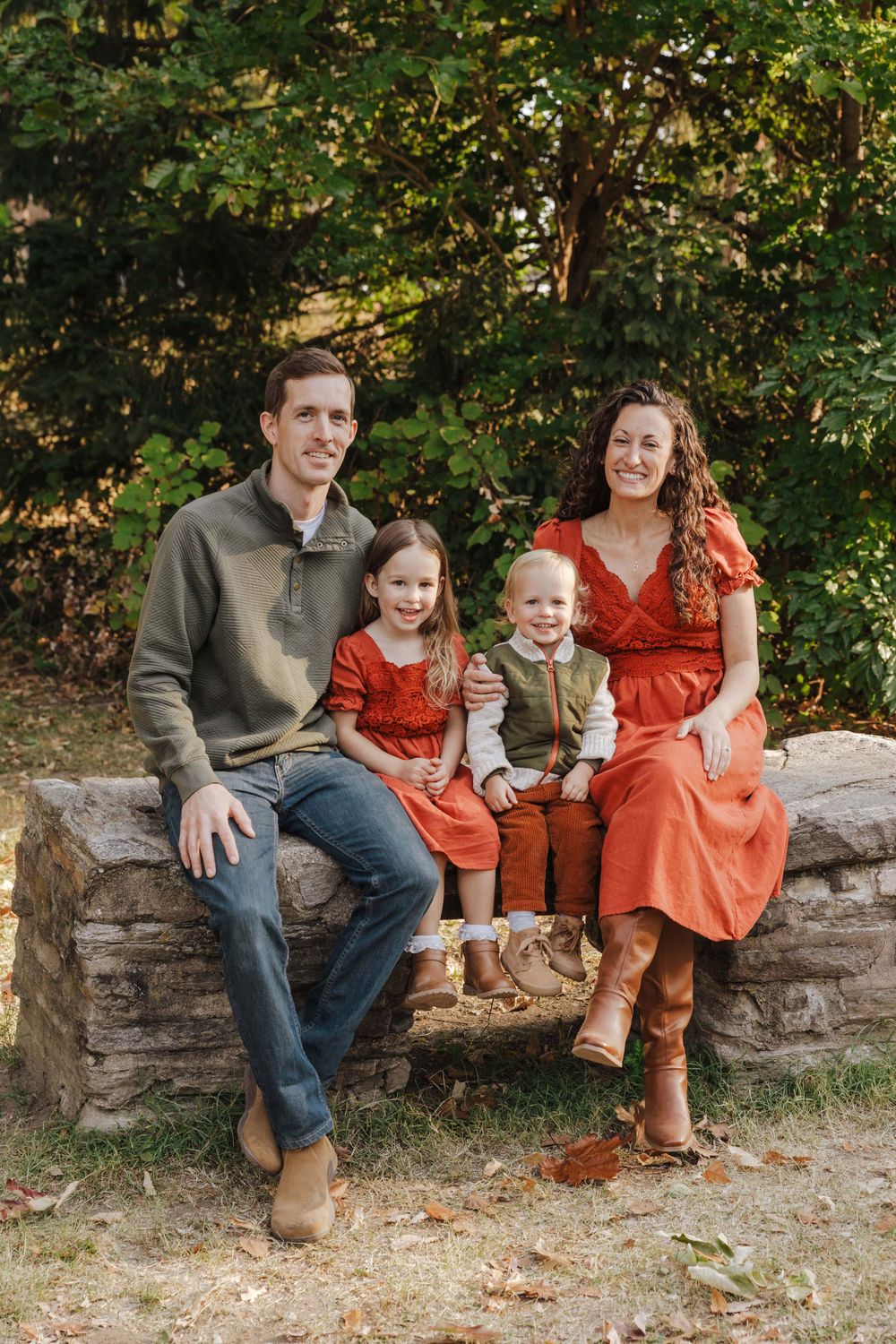 A family wearing coordinating orange and neutral tones sits together on a stone wall in an autumn outdoor setting.
