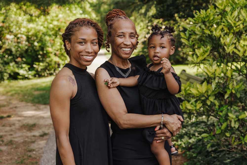 Three people wearing black outfits smile warmly together outdoors on a sunny path with greenery in the background.