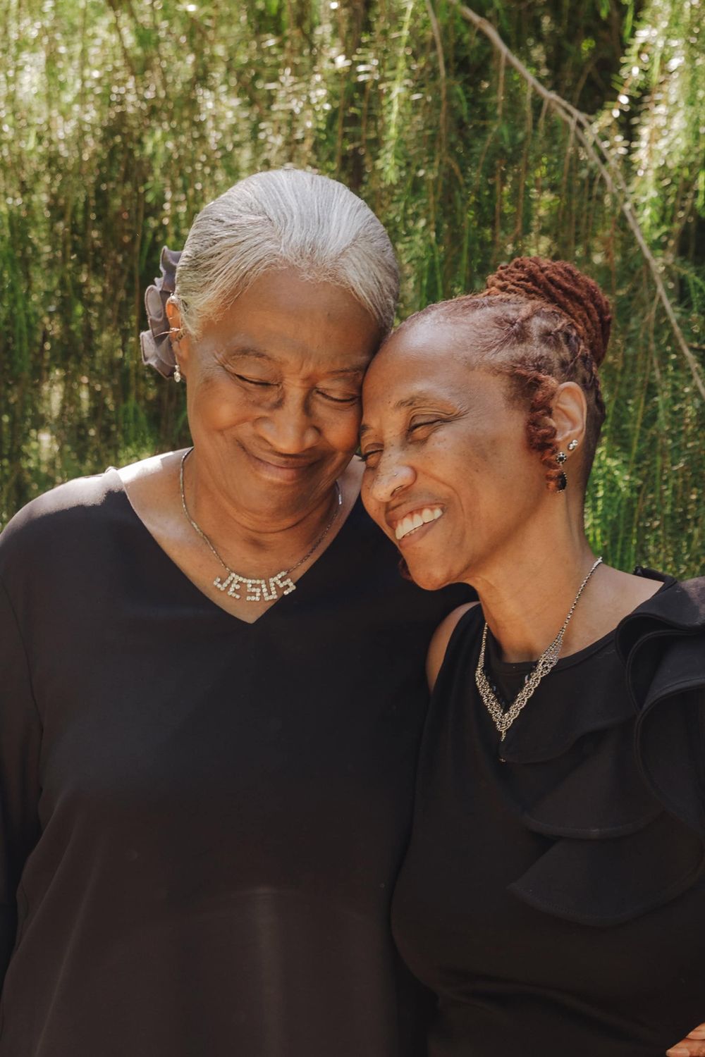 Two generations share a tender moment of joy while wearing black tops and delicate necklaces outdoors.