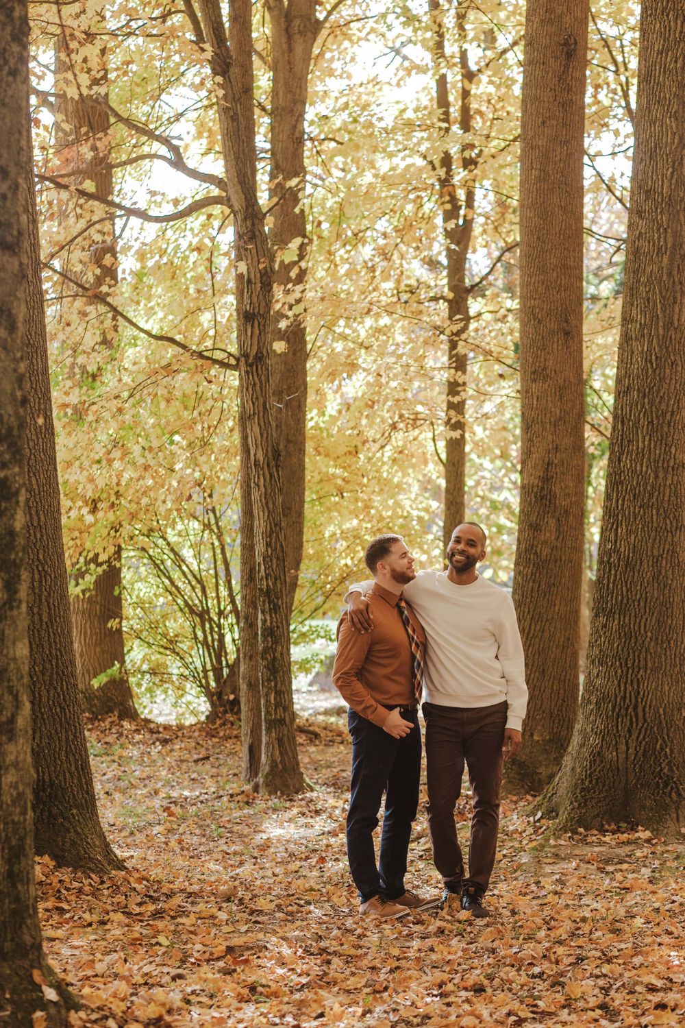 A couple walks together on a leaf-covered forest path during autumn, surrounded by tall trees in warm golden light.