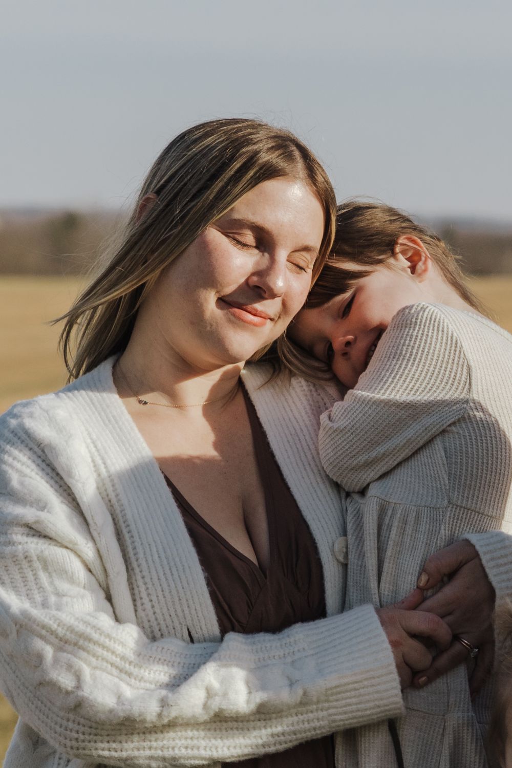 Two people in white sweaters share a tender moment outdoors with soft sunlight and a blurred field in the background.
