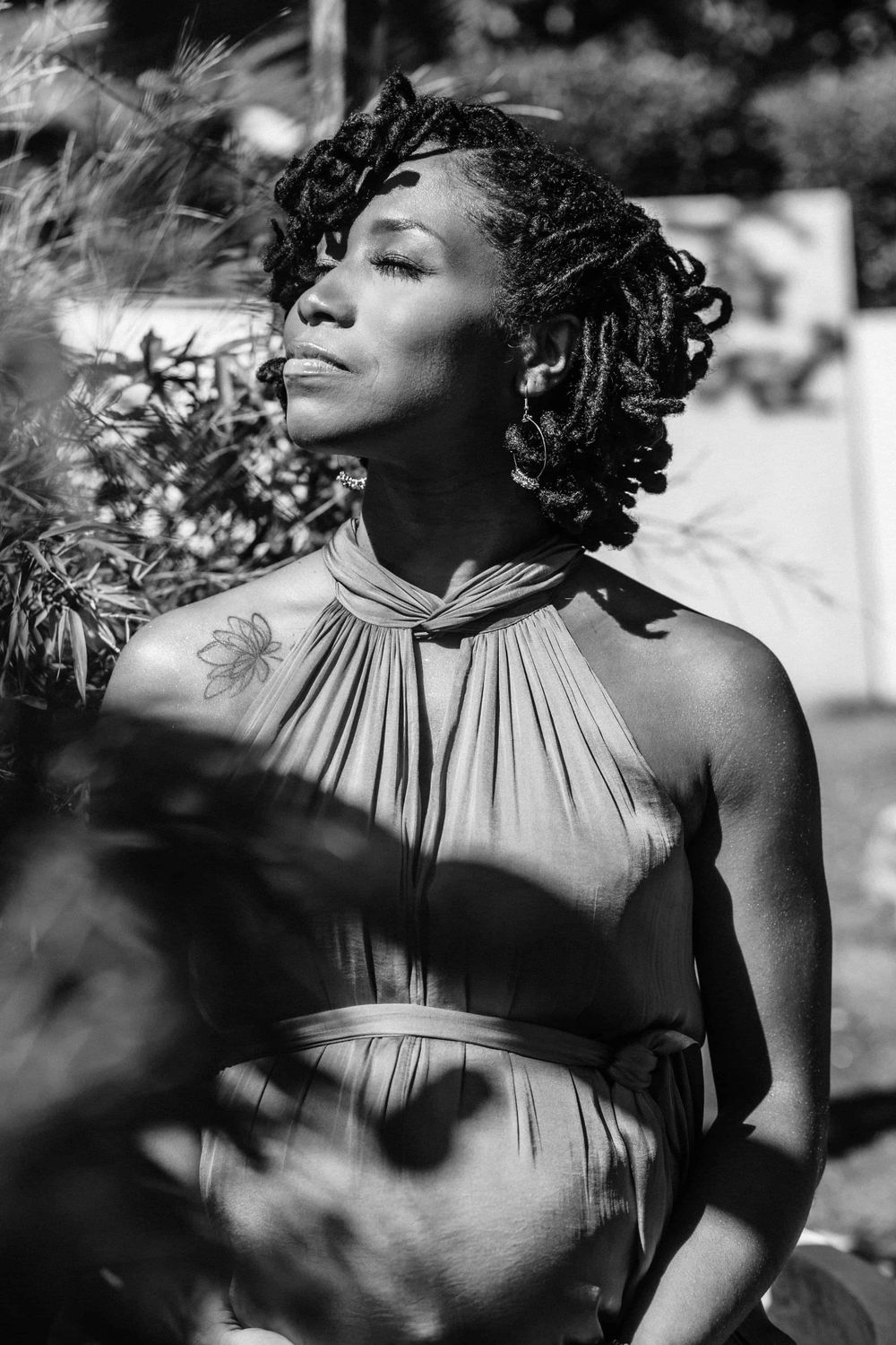 A black and white artistic portrait showing a person in a halter dress posing outdoors in dappled sunlight.