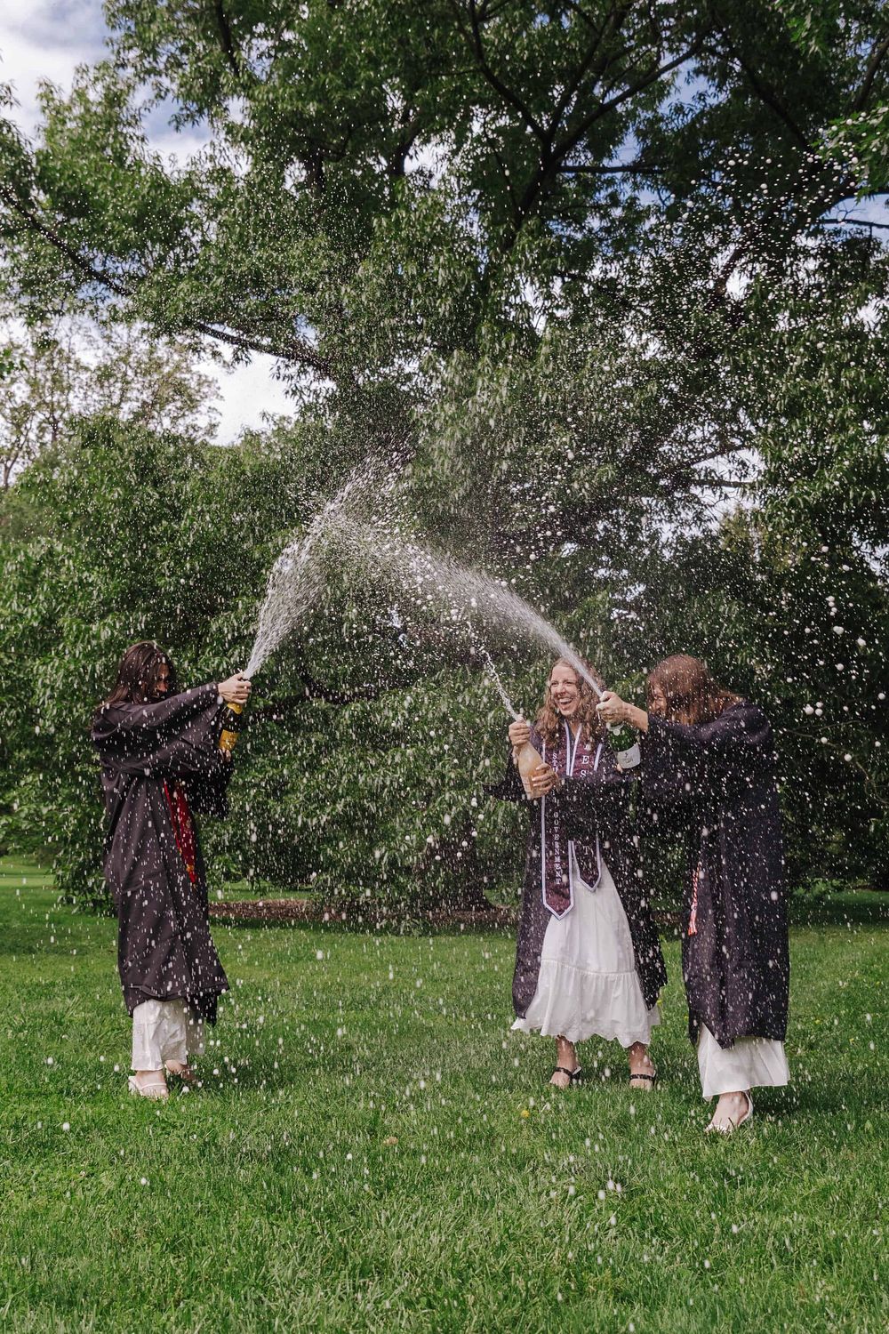 Graduating seniors celebrate with champagne spraying in a lush green outdoor setting.