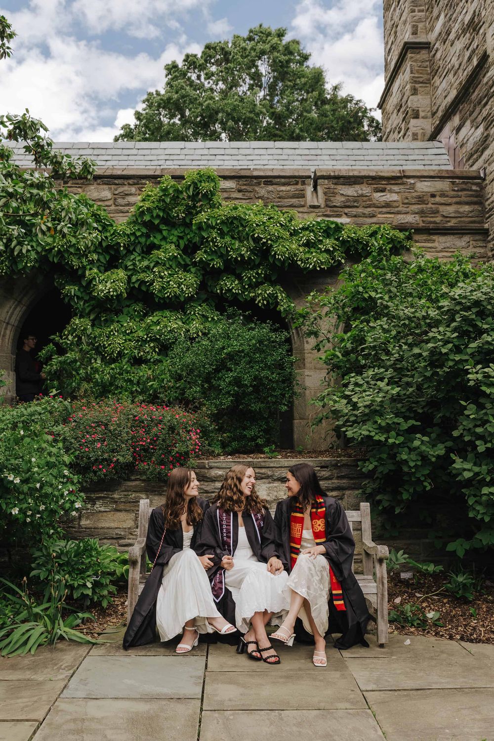 Three graduating seniors sit together on stone steps surrounded by lush green foliage on a sunny day.