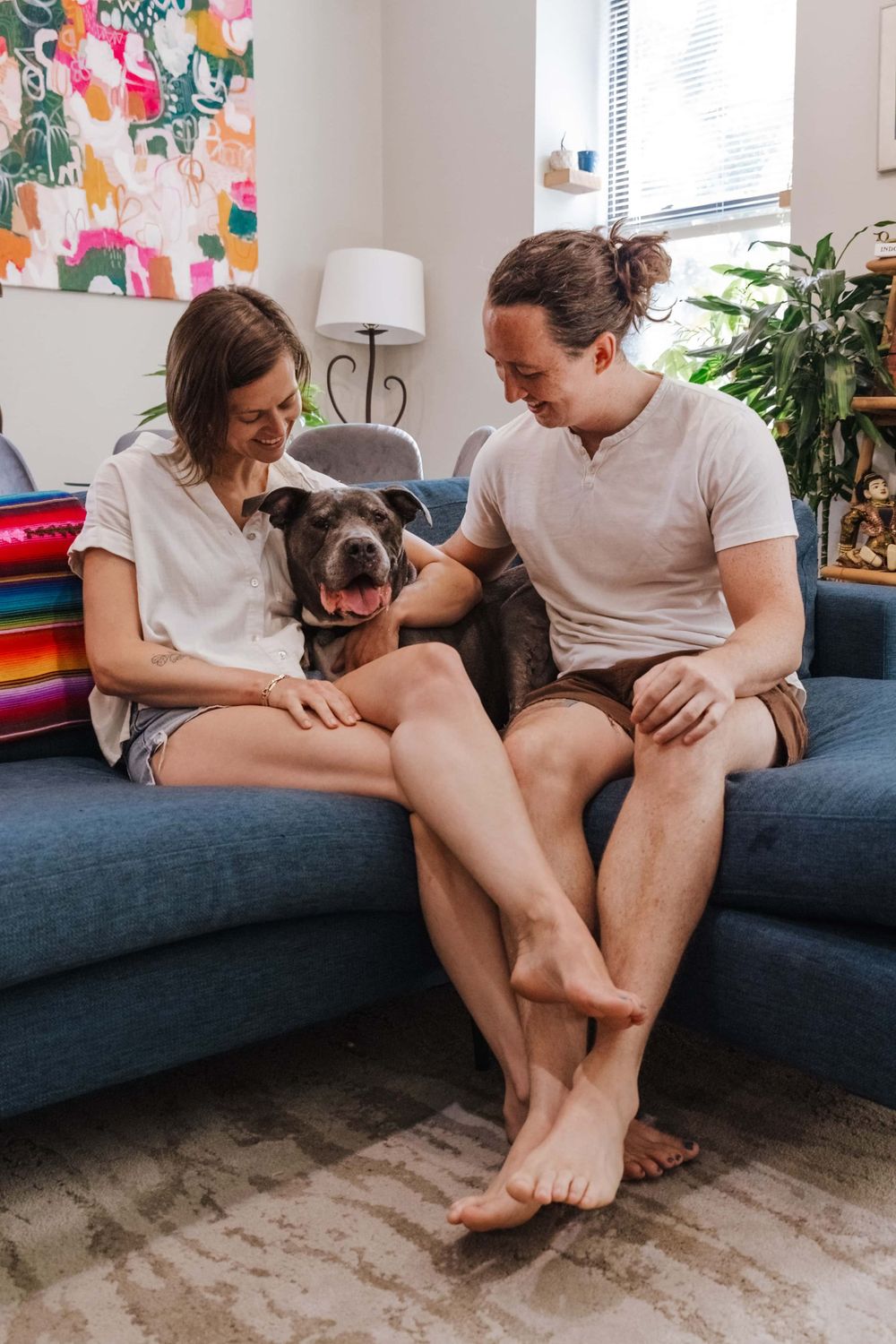 Couple relaxes on blue sofa with their gray dog while enjoying a bright, plant-filled living room with colorful wall art.
