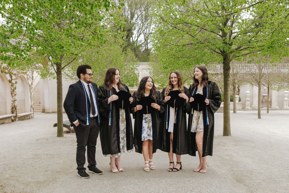 A group of graduates in black and white attire stand together on a tree-lined path in an outdoor setting.
