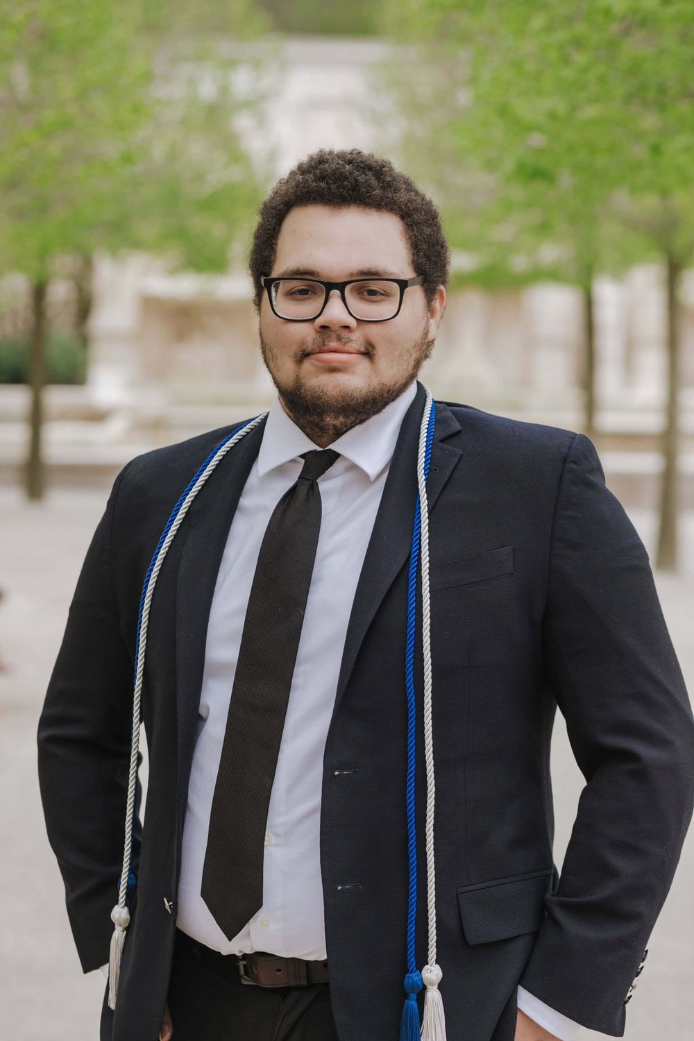 Graduate wearing formal black suit with glasses and blue graduation stole stands outdoors on campus path.