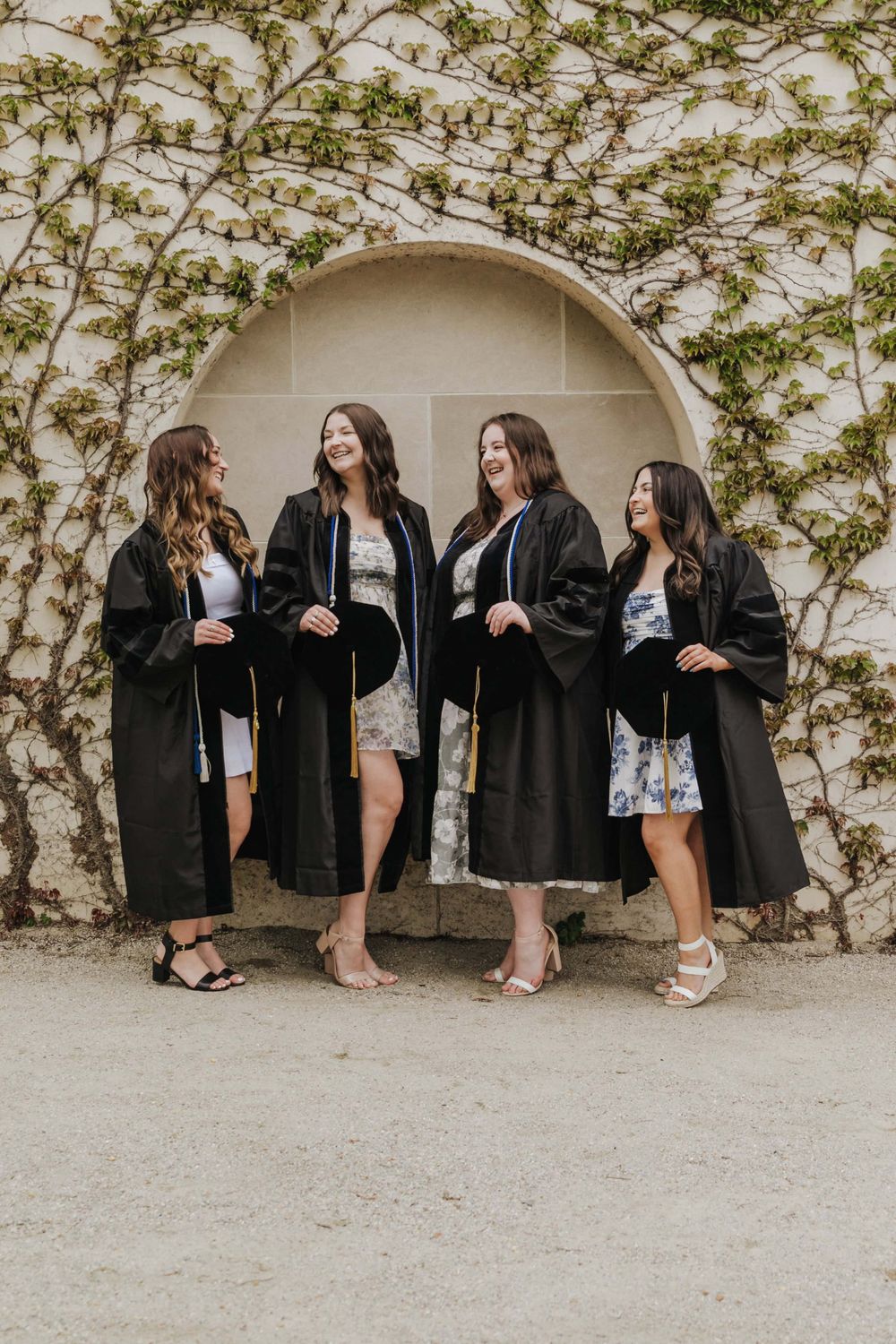 Group of graduates wearing black graduation robes standing together under an archway with climbing vines on the wall.