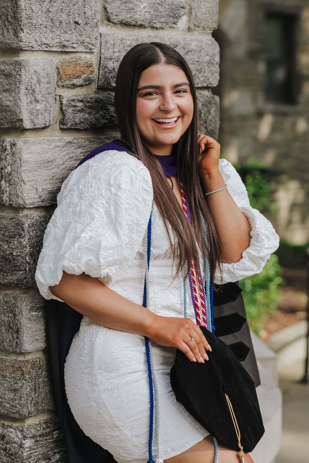 Person in white summer dress with puff sleeves leans against stone wall while smiling and holding black bag.