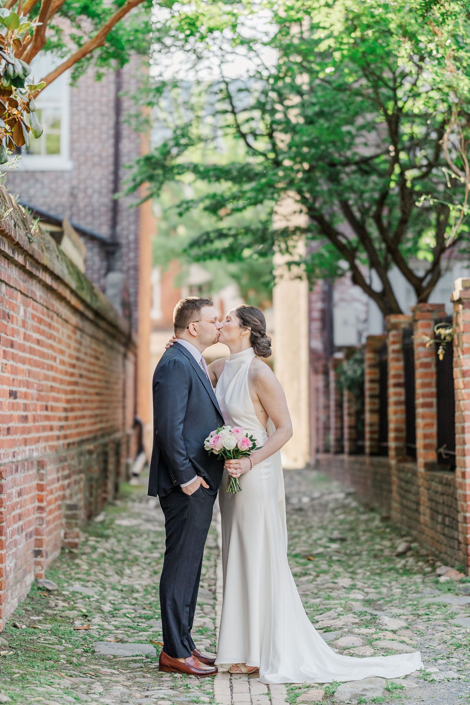 A bride and groom share a romantic kiss in a charming brick alleyway, the bride holding a pink bouquet in her elegant white gown.
