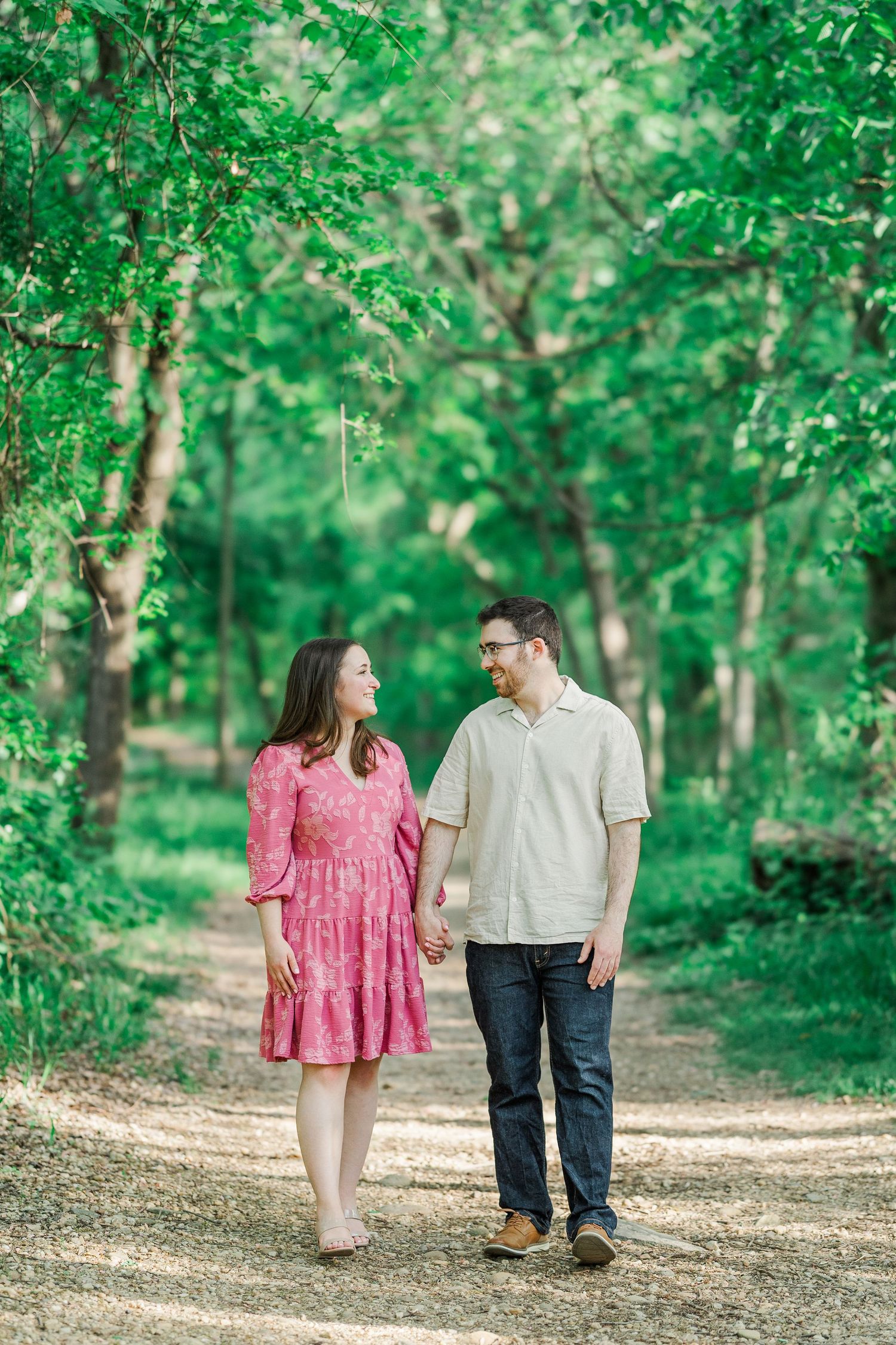A couple walks hand-in-hand on a forest path, smiling at each other, woman in pink dress, man in casual attire.