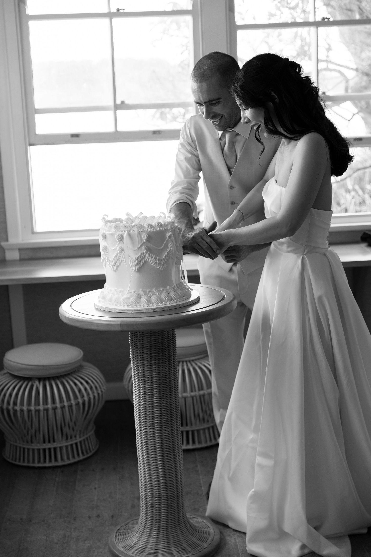 Black and white photo of a bride and groom cutting their wedding cake together near a sunlit window.