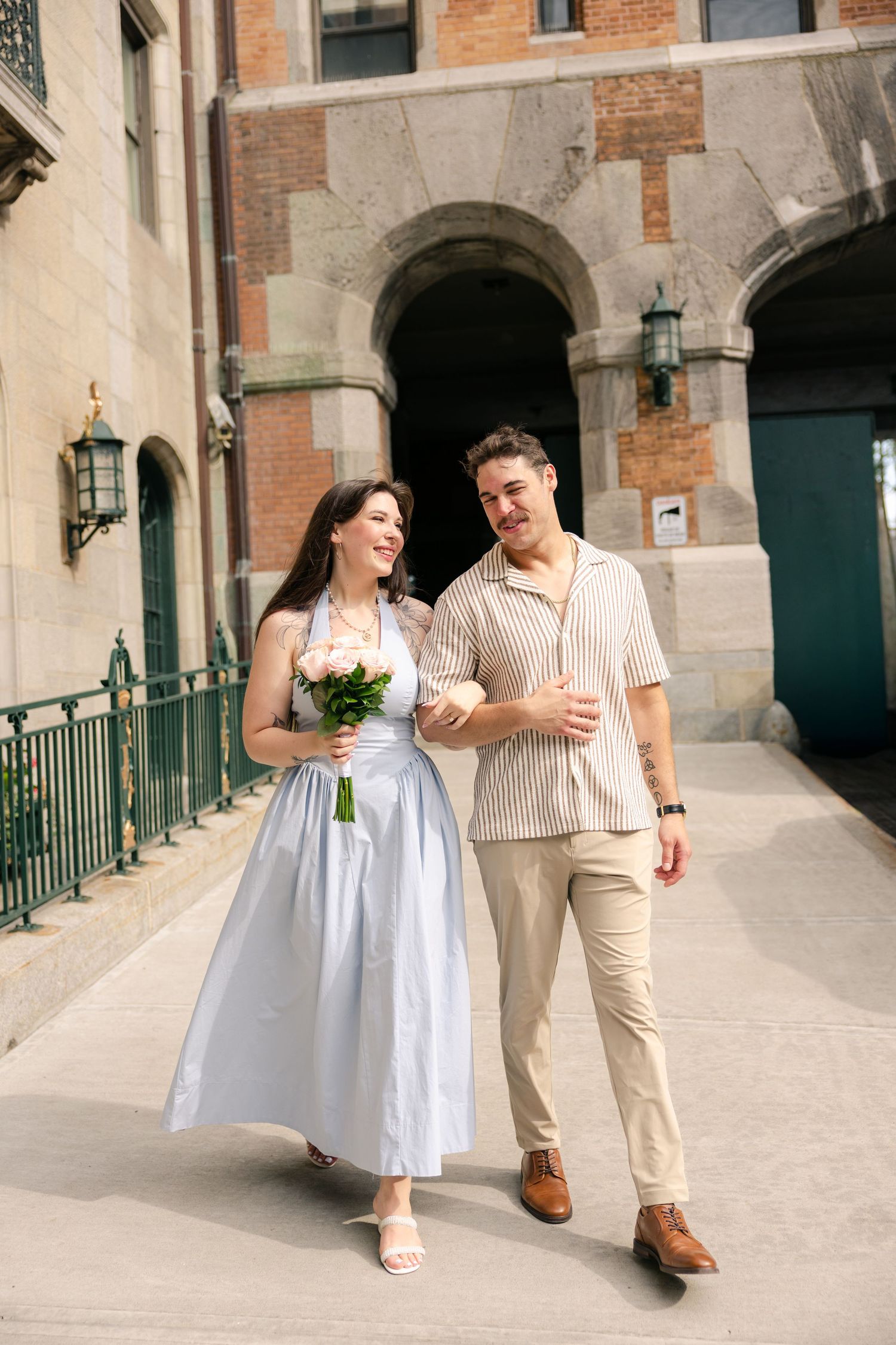 Happy couple walking outdoors, woman in white dress holding bouquet, man in beige shirt, near historic building.