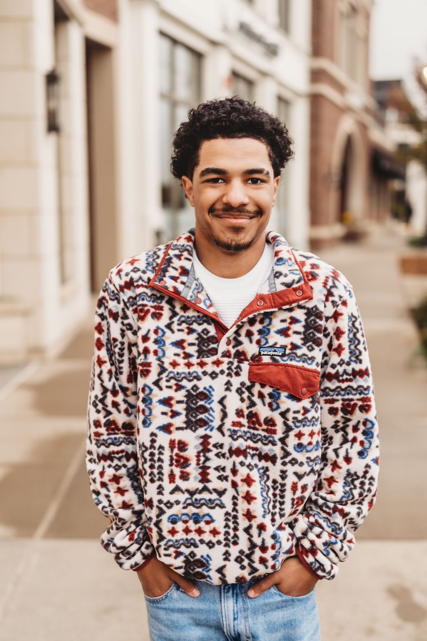 Young man smiling outdoors in a patterned Patagonia fleece pullover and jeans, standing on a city sidewalk.