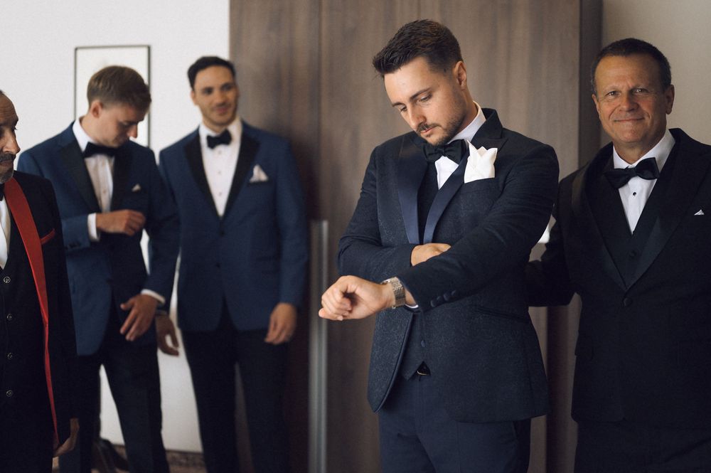 Groom checks his watch while groomsmen in navy tuxedos prepare together before a wedding ceremony.