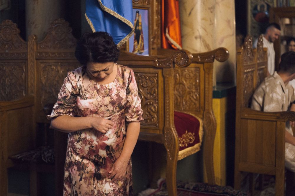 A woman in a floral qipao dress bows her head in prayer inside an ornate wooden church interior.