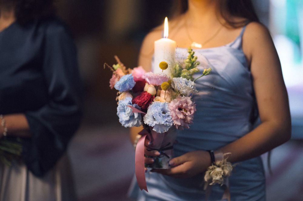 Bridesmaid in blue dress holding floral bouquet with lit white candle at wedding ceremony.