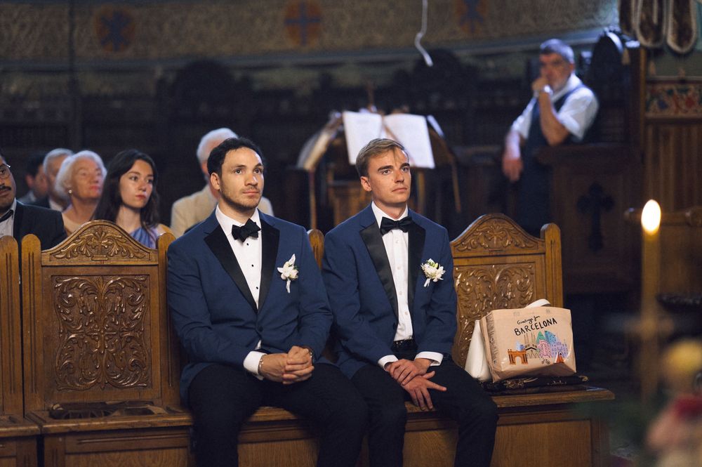 Two men in navy tuxedos with white boutonnieres seated in ornate wooden church pews during a wedding ceremony.