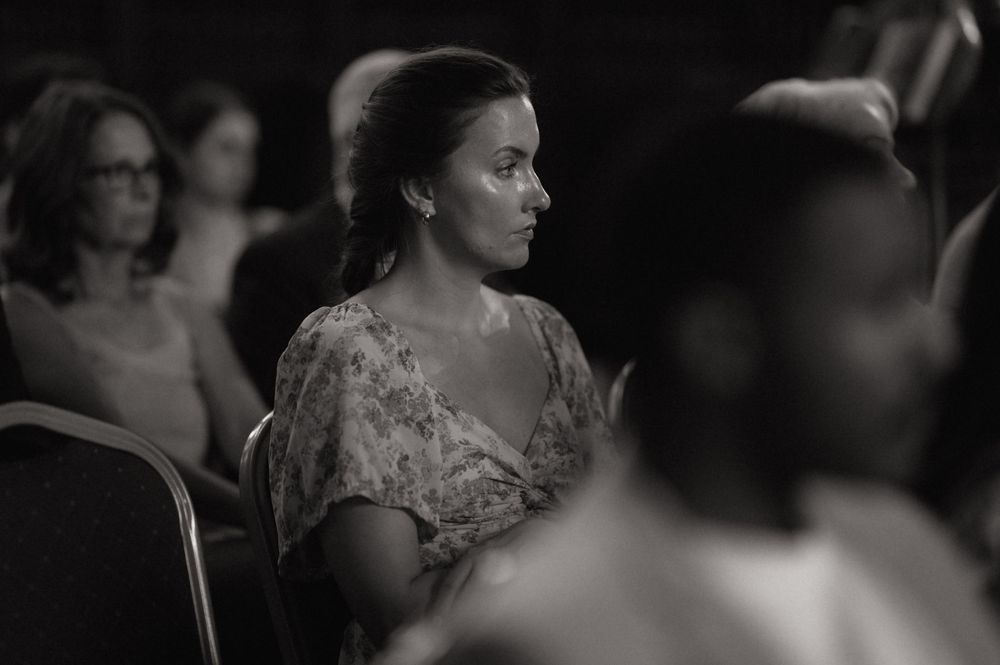 Black and white photo of woman in floral dress sitting thoughtfully in audience at event.