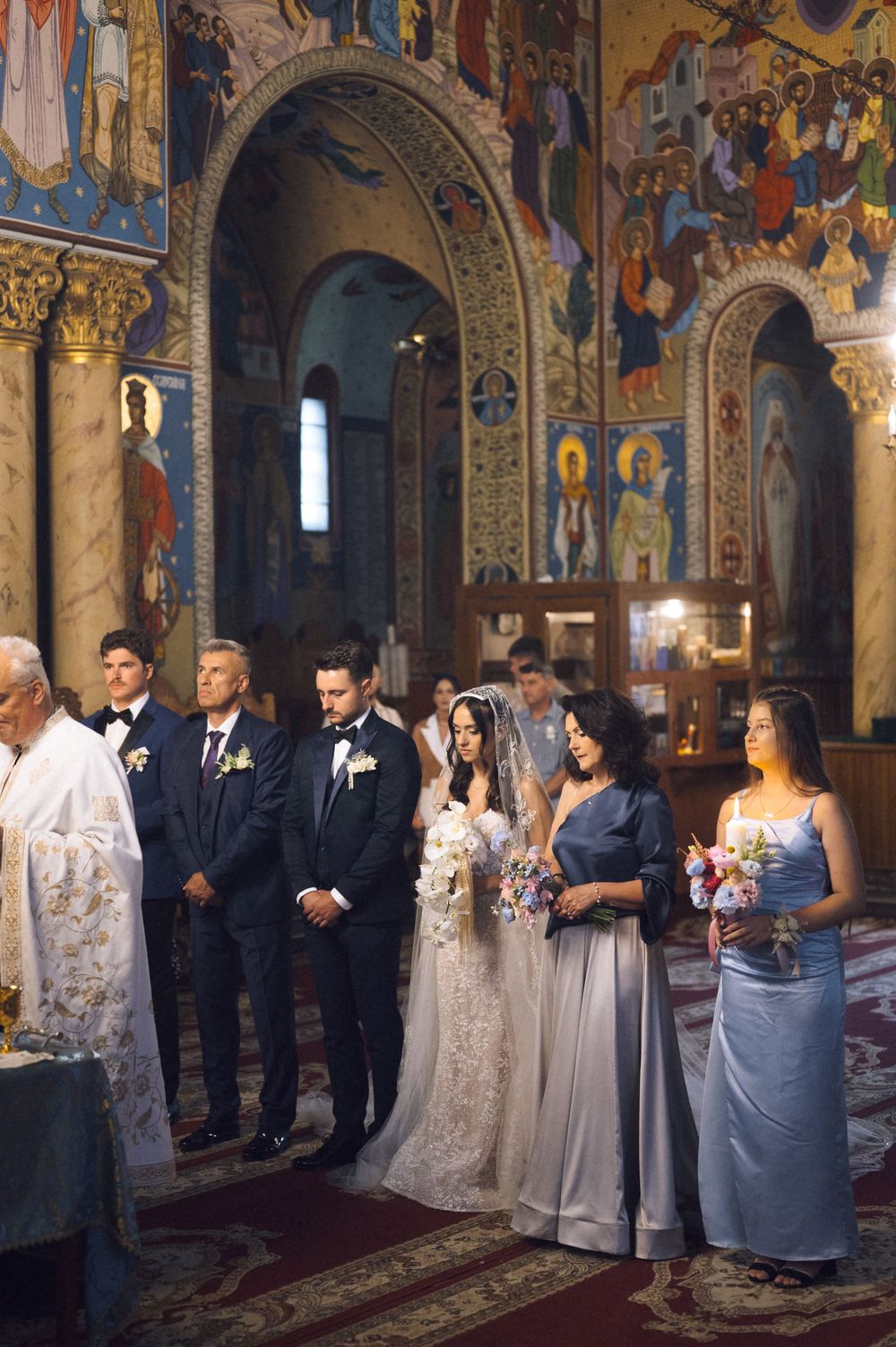 Wedding ceremony in ornate Orthodox church with bride in veil, groom in tuxedo, and guests amid colorful frescoes.