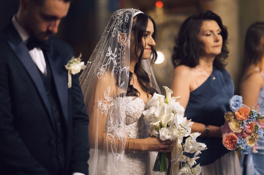 Bride in lace veil holding white calla lily bouquet at wedding ceremony with guests in background.