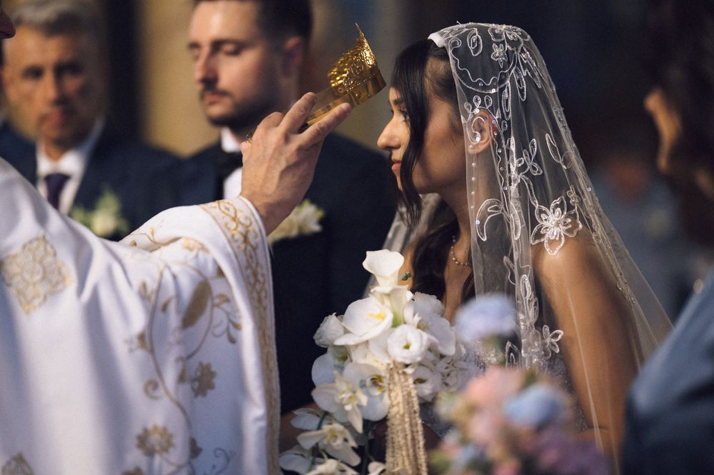 Bride in lace veil receives communion from priest during traditional church wedding ceremony.