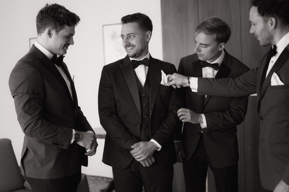 Four groomsmen in black tuxedos sharing a moment while getting ready for a wedding ceremony.