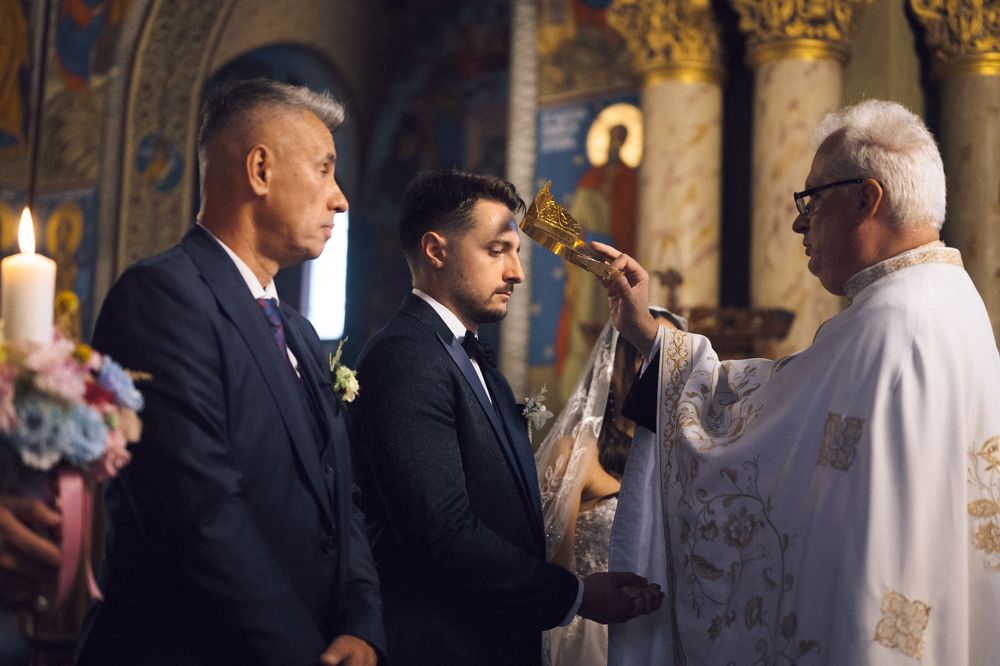 Priest performing a religious blessing ceremony on two men in suits inside an ornate Orthodox church.