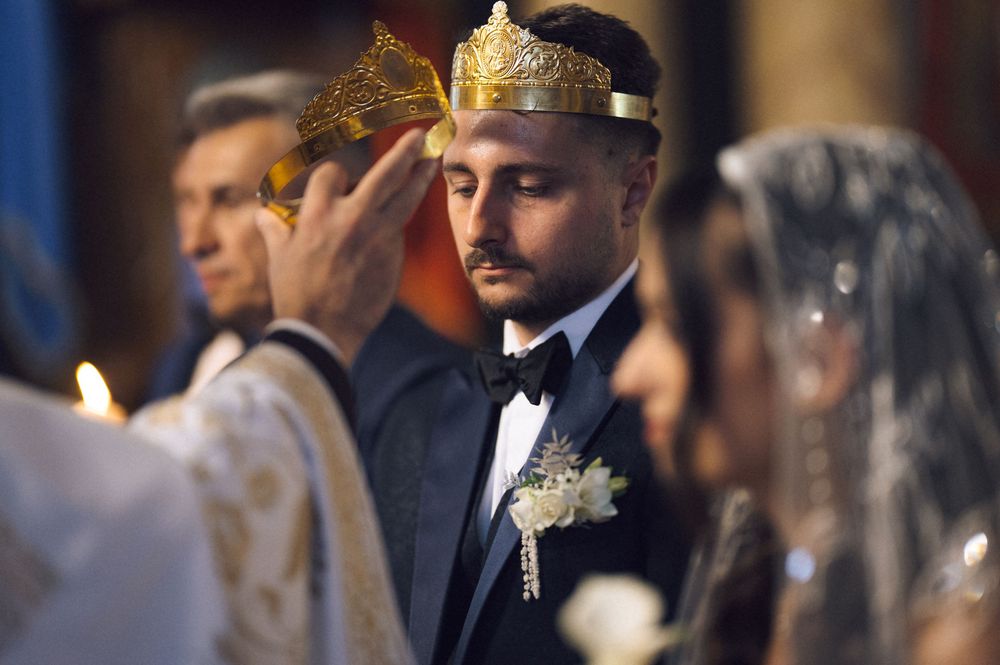 Groom wearing gold crown during Orthodox wedding ceremony with priest and veiled bride in church.