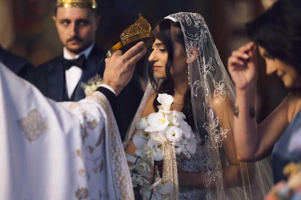 Orthodox wedding ceremony with bride in lace veil receiving crown from priest, groom in tuxedo standing behind.