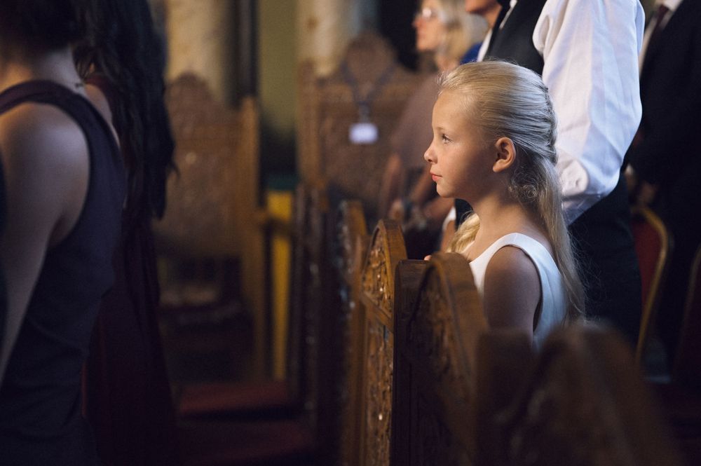 Young blonde girl in white dress standing attentively in wooden church pews during a religious service.