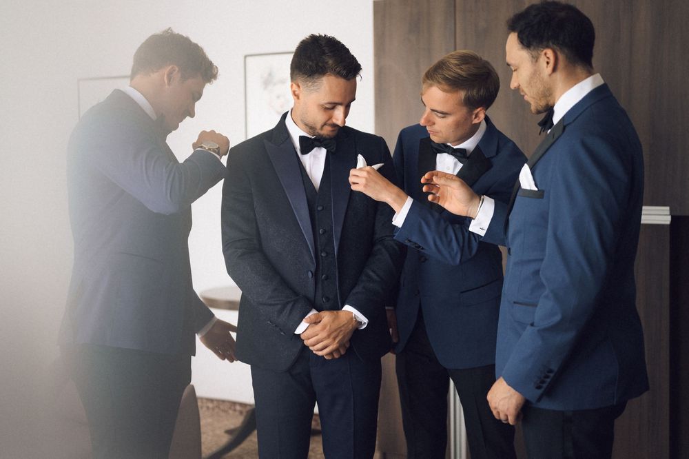 Four men in formal suits and bow ties helping each other get dressed for a wedding ceremony.