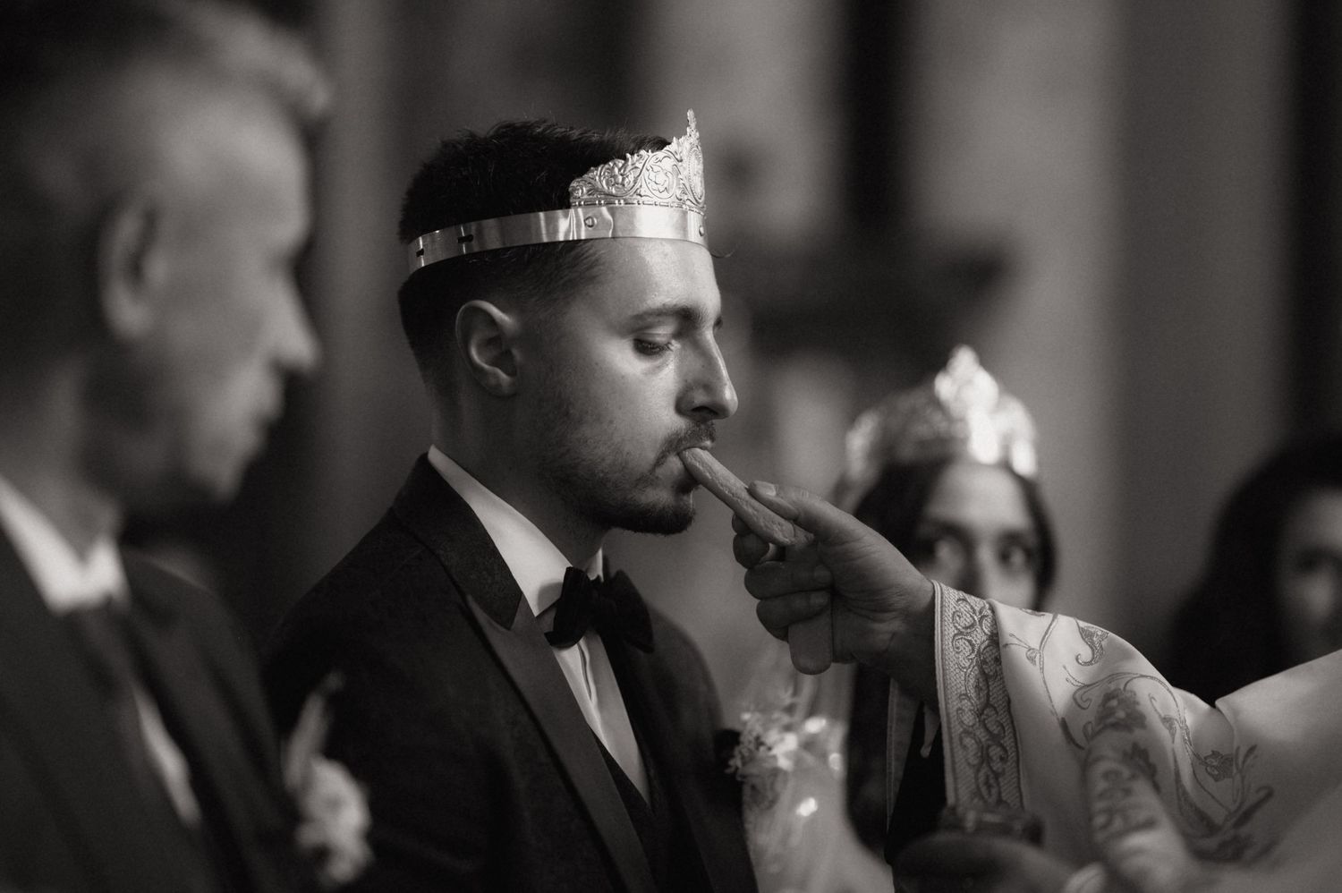 Black and white wedding photo of groom wearing crown kissing bride's hand at ceremony.