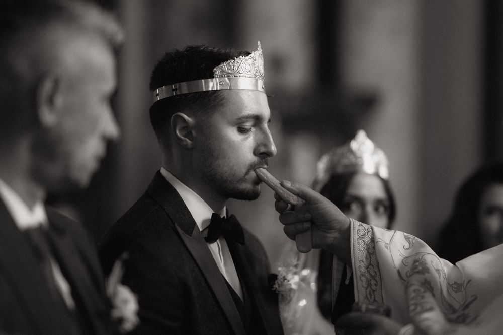 Black and white wedding photo of groom wearing crown kissing bride's hand at ceremony.