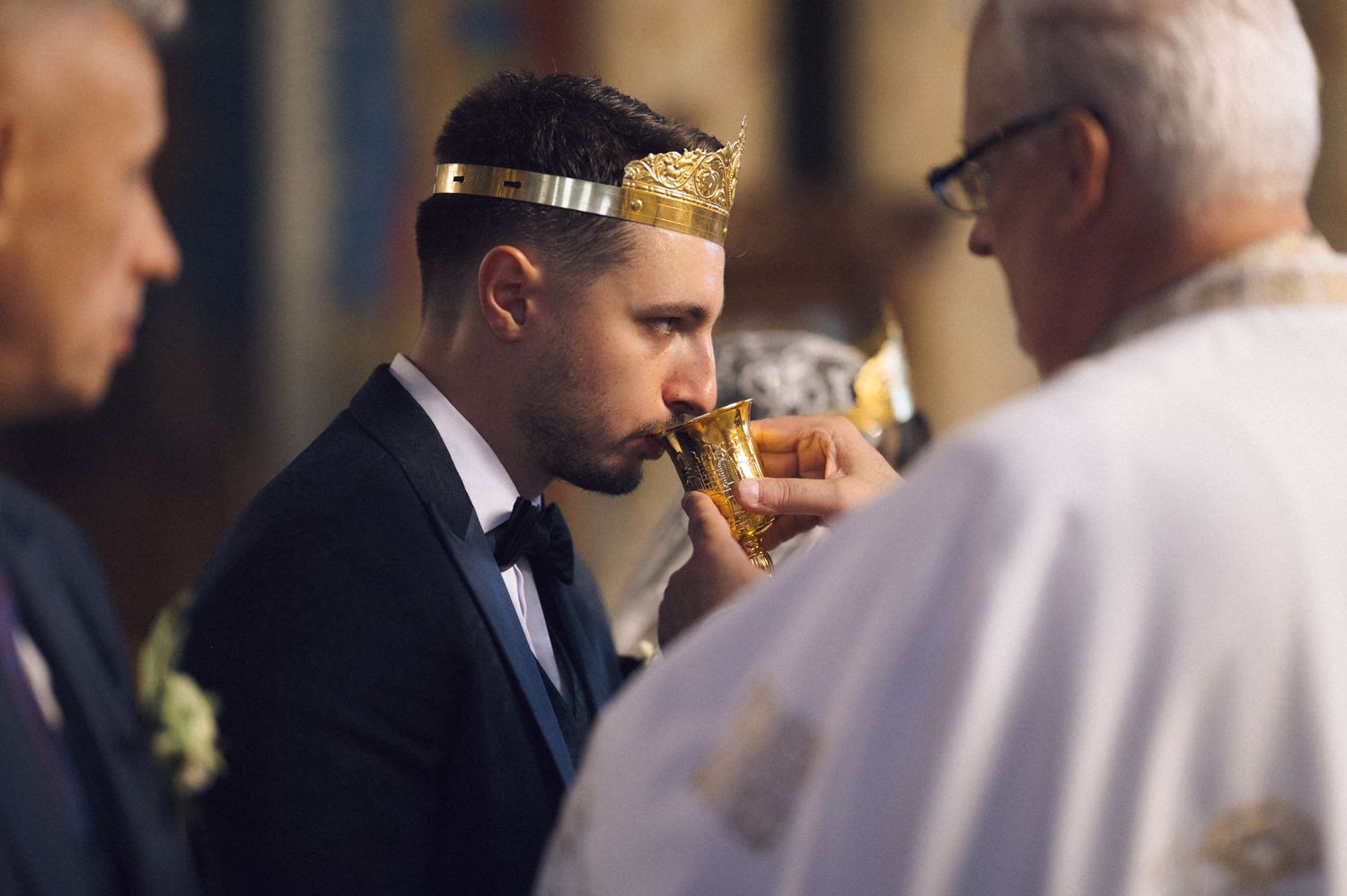 Groom wearing gold crown drinks from chalice during religious wedding ceremony officiated by priest.