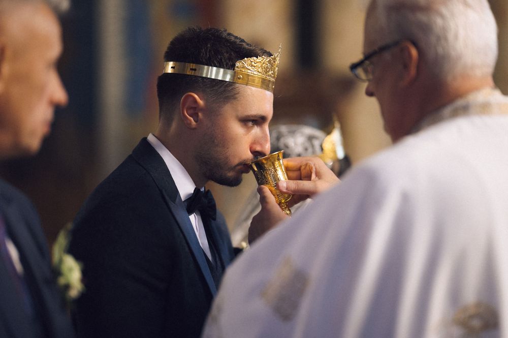 Groom wearing gold crown drinks from chalice during religious wedding ceremony officiated by priest.