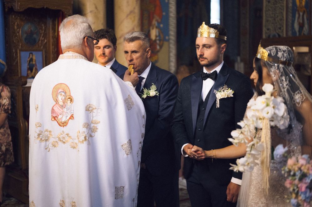 Orthodox wedding ceremony with groom in suit wearing crown, bride in veil, and priest in white vestments at church altar.